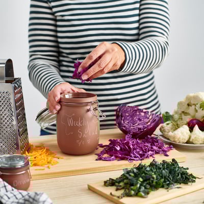 Person preparing salad with KORKEN jar, shredding cabbage on wooden board, surrounded by vegetables.