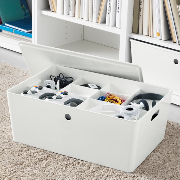 White organizer box with multiple compartments holding game controllers and cables, placed on rug near bookshelf.
