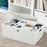 White organizer box with multiple compartments holding game controllers and cables, placed on rug near bookshelf.