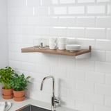 Kitchen shelf with cups, glasses, and plates, mounted to a tiled wall. Two potted herbs sit on a counter below a chrome faucet.