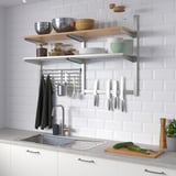 Stainless steel shelves above sink with bowls, pans, ingredients, utensils & knives. Cutting board with cucumbers, plant, soap & towels.