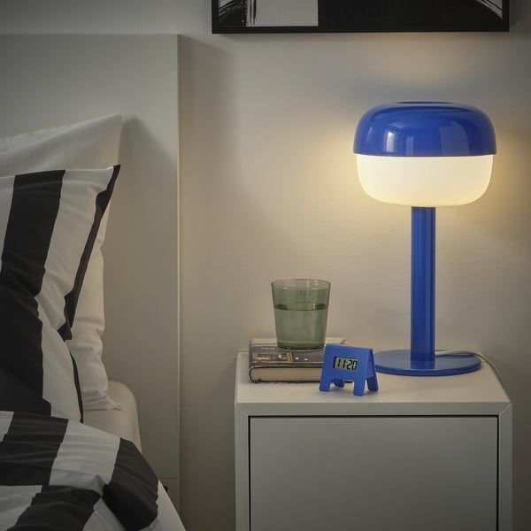 Bedside scene with bright blue KUPONG alarm next to lamp and glass. Clock is small, fitting nightstand near bed.