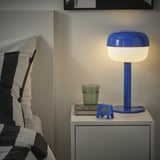Bedside scene with bright blue KUPONG alarm next to lamp and glass. Clock is small, fitting nightstand near bed.
