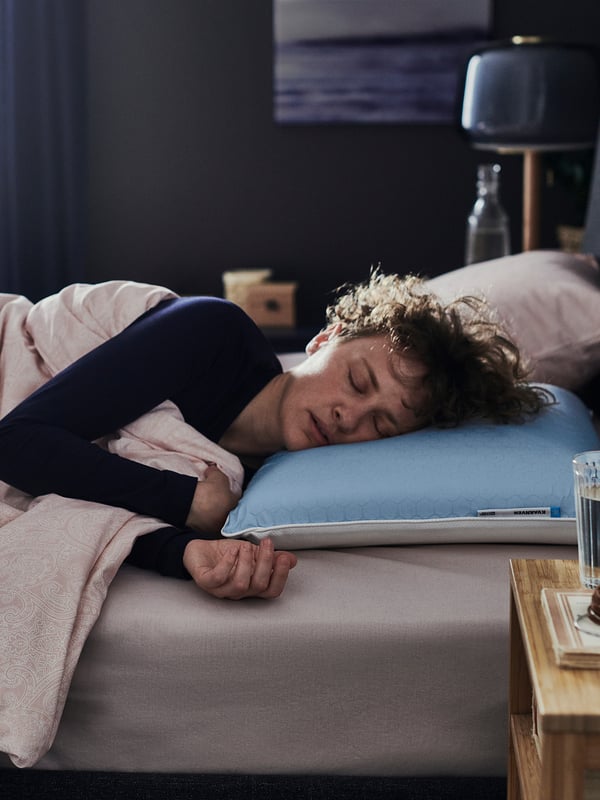 A person sleeps on a bed with a blue KVARNVEN honeycomb-patterned pillow and a wooden nightstand with a glass of water.