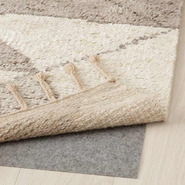 Light cream, hand-knotted area rug with fringe and geometric pattern. Underlay visible, placed on wood floor.