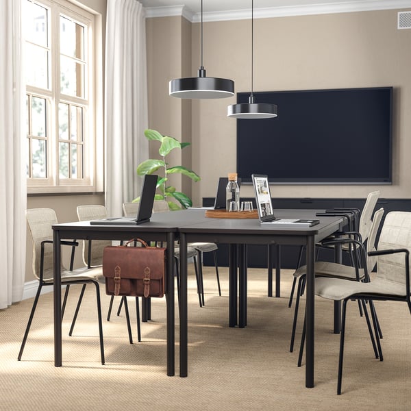 Modern conference room with dark tables, chairs, tv, and hanging lamps. Natural light and plant decor.
