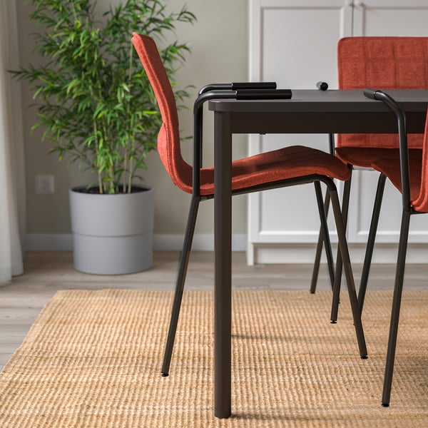 Stackable conference chairs with armrests and lumbar support sit on a woven rug under a potted plant, showcasing a clean, organized meeting space.