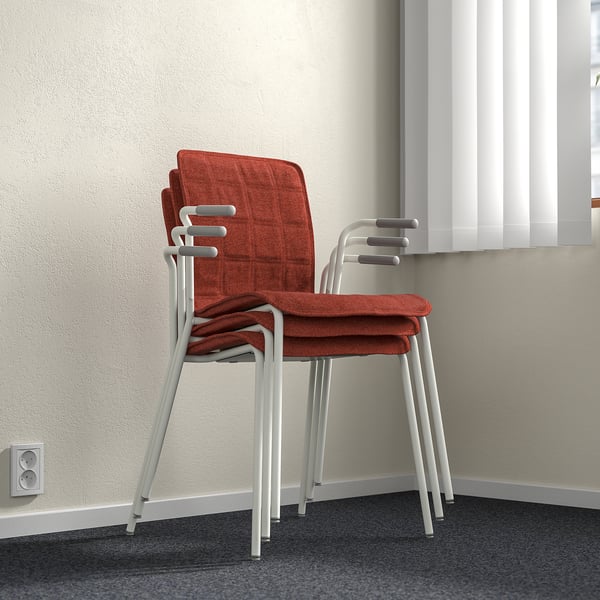 Stacked chairs with orange fabric and white metal frames against a wall with blinds.