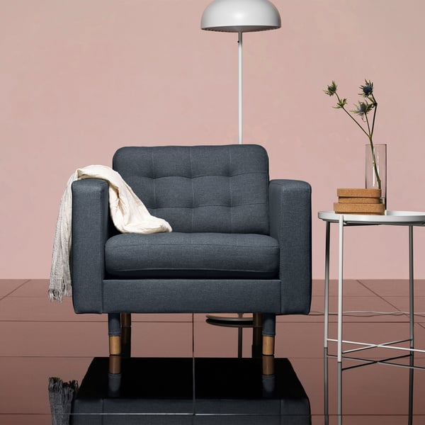 Cozy grey armchair with wood legs beside small table, lamp, and flowers. Soft, modern design.