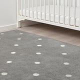 Childs room with white crib, grey rug with white dots, placed on wooden floor.