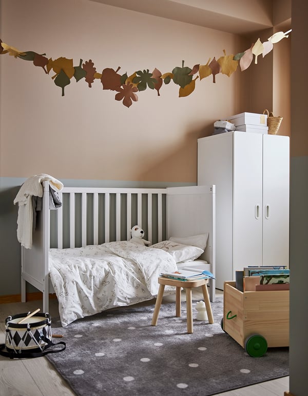 Childs room with white bed, grey dot rug, wooden toy box, drum, white cabinet, and leaf decoration on wall.