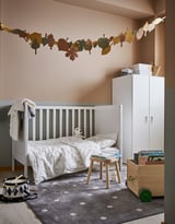 Childs room with white bed, grey dot rug, wooden toy box, drum, white cabinet, and leaf decoration on wall.