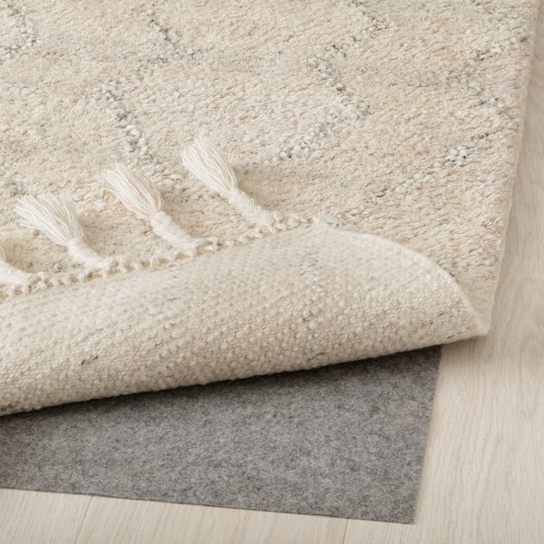 A fluffy, cream-colored wool rug with fringed edges, placed on a gray rug underlay for stability and comfort.