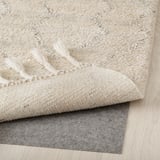 A fluffy, cream-colored wool rug with fringed edges, placed on a gray rug underlay for stability and comfort.