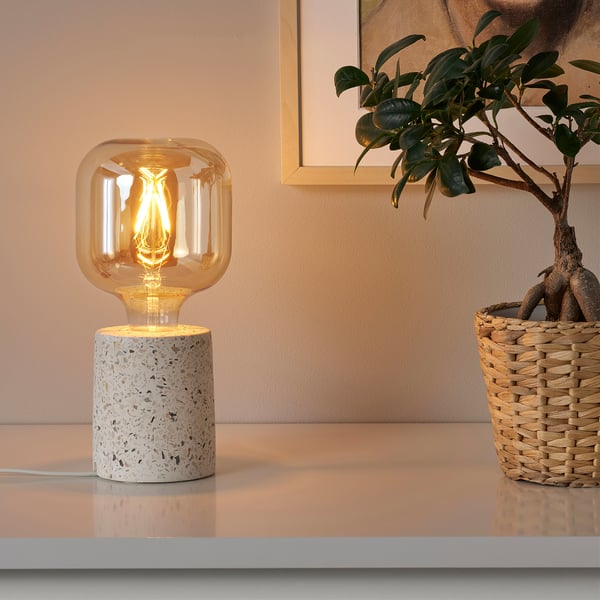 Table lamp with warm lightbulb and woven plant holder on shelf.