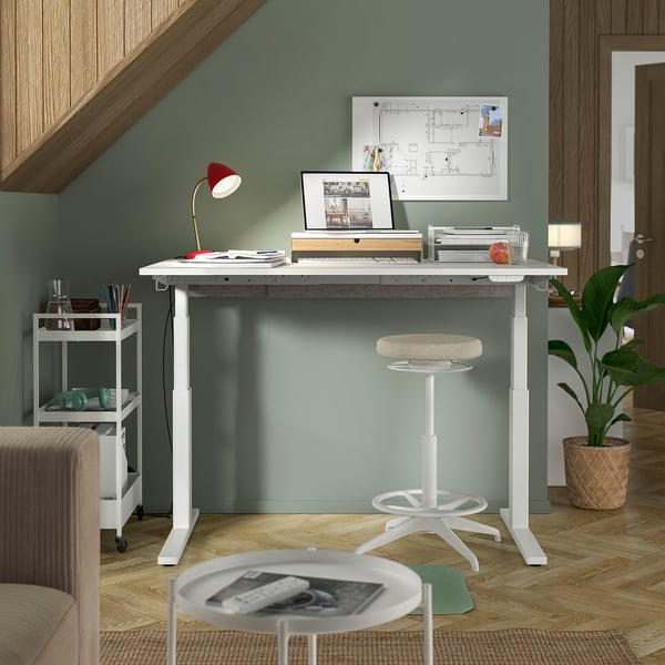 A modern home office with a white adjustable desk, red lamp, laptop, and a beige stool. Green accent wall.