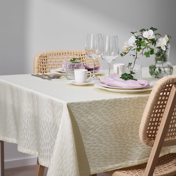 Table set with shimmering white tablecloth, trendy woven chairs, plates, glasses, cups, cutlery, and pink napkins with flowers.