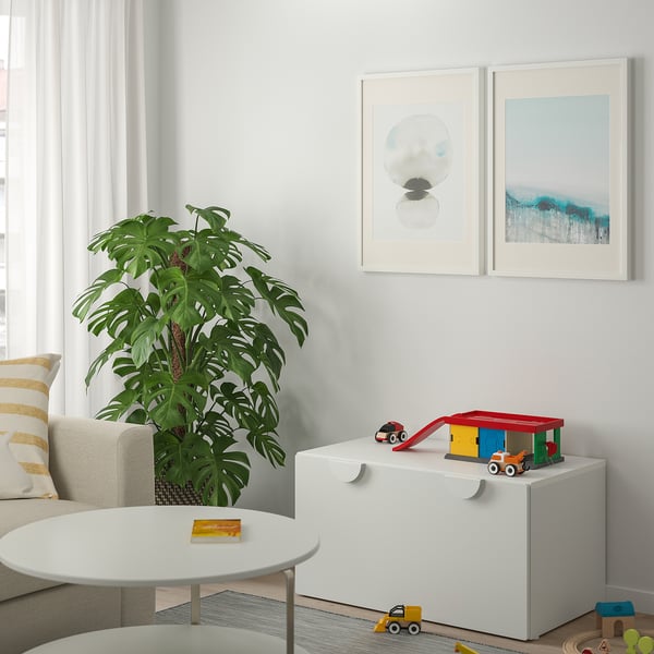 Bright toy garage playset on white storage box amidst living room, near potted plant and art frames.