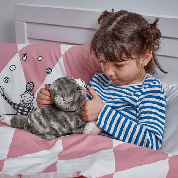 A young girl embraces a plush gray kitten toy while lying in bed, likely seeking comfort and companionship.