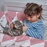 A young girl embraces a plush gray kitten toy while lying in bed, likely seeking comfort and companionship.