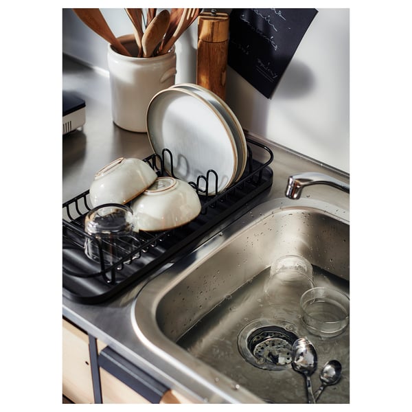 Black LILLHAVET dish rack with plates, bowls, cups, and cutlery. Stainless steel sink, wooden utensils, and white countertop.