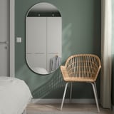 Modern bedroom: green wall, large white oval mirror, wicker chair, white curtains, bed. Mirror larger than chair.