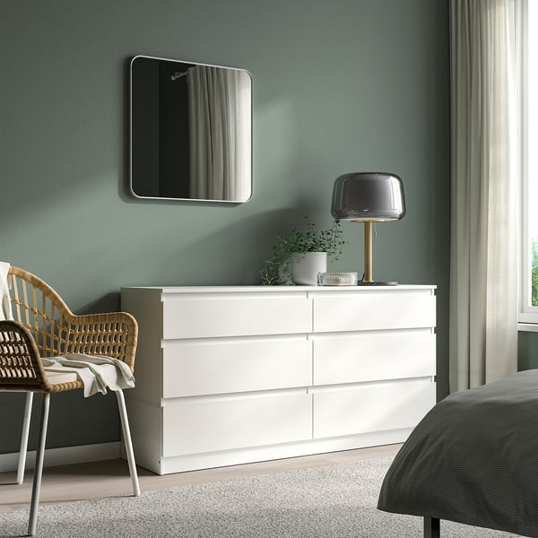 Modern bedroom with LINDBYN mirror. White drawers, grey bedding, wicker chair, black lamp. Large mirror above dresser.