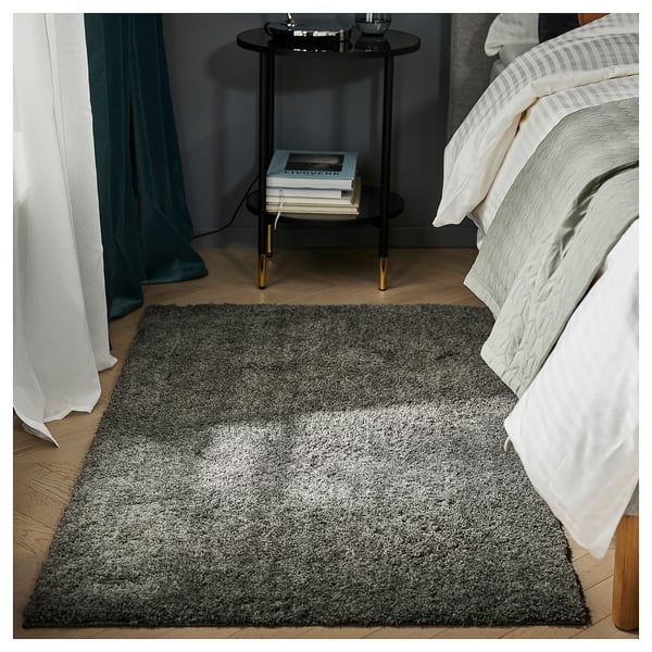 Bedroom with grey LINDKNUD rug, white bedding, black nightstand, teal curtains, and stack of books.