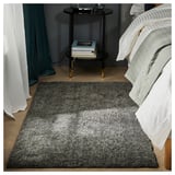 Bedroom with grey LINDKNUD rug, white bedding, black nightstand, teal curtains, and stack of books.