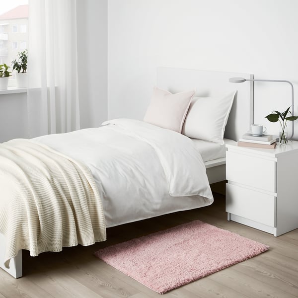 Minimalist bedroom with white bed, pink LINDKNUD rug, and table. Recycled polyester rug adds soft touch.