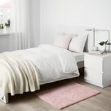 Minimalist bedroom with white bed, pink LINDKNUD rug, and table. Recycled polyester rug adds soft touch.