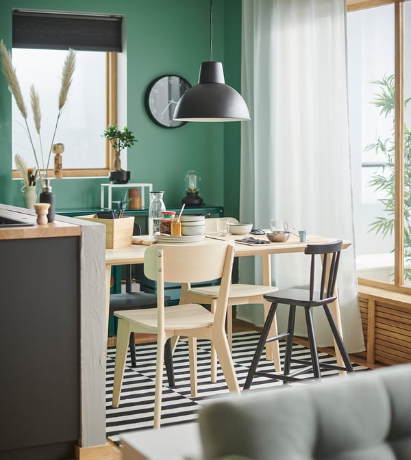 Modern dining area with green walls, black ceiling light, wooden table, four light wood chairs, striped rug, and large window with white curtains.