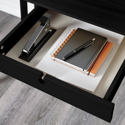 Black desk drawer with notebook, pen, binder clip, and stapler, showcasing organized workspace.