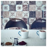 Two white cups with blue handles on a table with decorative tiles and backs of chairs behind, accompanied by tea infusers and heaps of spices.