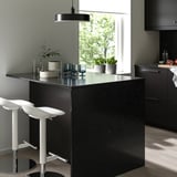 Modern kitchen with black glass countertop, stools, blue glasses, plants, light wood floors, dark cabinets, and large window.