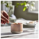 Hand lighting brown ceramic LUGNARE candle on table with greenery.