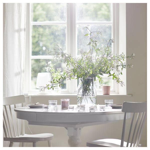 Bright dining area with white round table, chairs, and flowers in a vase by a large window.