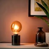 A small table lamp with a warm-colored light bulb on a dark countertop, beside a framed plant picture and decorative bottles.