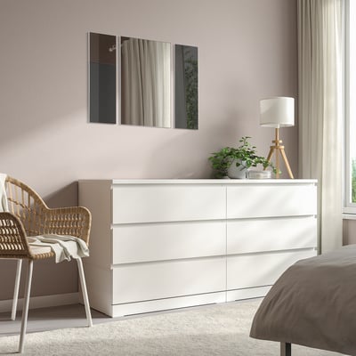 A modern bedroom with MADESJÖ white dresser, tall and rectangular, topped with table lamp and plant. Mirror and wicker chair beside it.