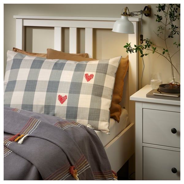 Cozy bedroom with plaid pillow, heart designs, wooden headboard, gray blanket, orange pillow, and nightstand with lamp, vase, glass.