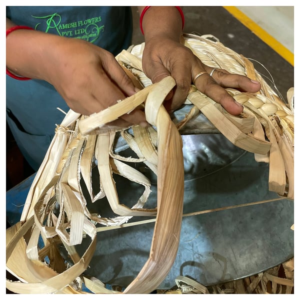 Person weaving long, thin, beige banana fibers on a blue metal surface, creating intricate patterns.