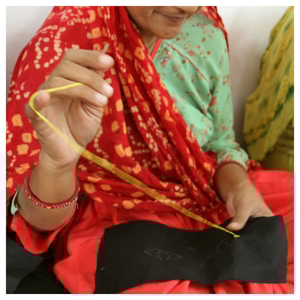A woman in a red and green patterned outfit is carefully sewing a yellow thread into a small black pouch.