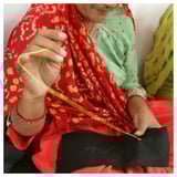 A woman in a red and green patterned outfit is carefully sewing a yellow thread into a small black pouch.
