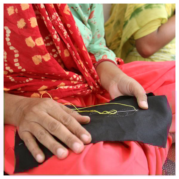 Two women in colorful traditional dresses sew, one with black fabric, the other with yellow thread, showcasing cultural artistry.