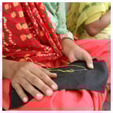 Two women in colorful traditional dresses sew, one with black fabric, the other with yellow thread, showcasing cultural artistry.