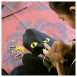 A person sews a black cat-shaped pouch on a pink floral cloth. The pouch yellow triangular ears and eyes, and a zipper mouth.