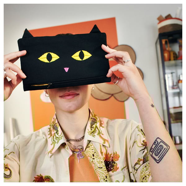 Person holding black cat-shaped pouch with glowing yellow eyes. Pouch has zipper and pointy ears. Person wears floral shirt and rings.