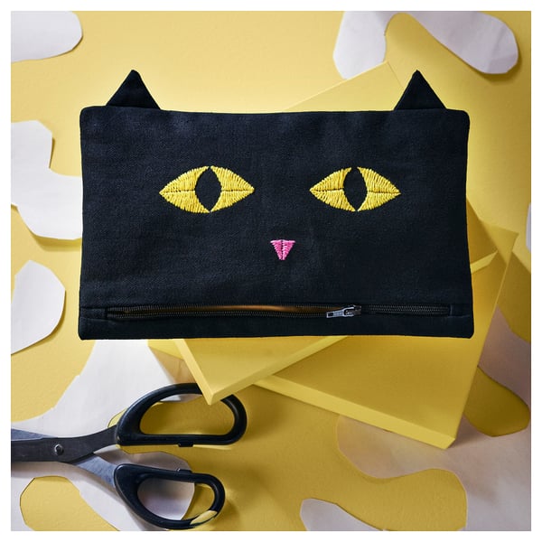 Black MÄVINN pouche with cat face design. Denim, rectangular with pointed ears, yellow fabrics, scissors.