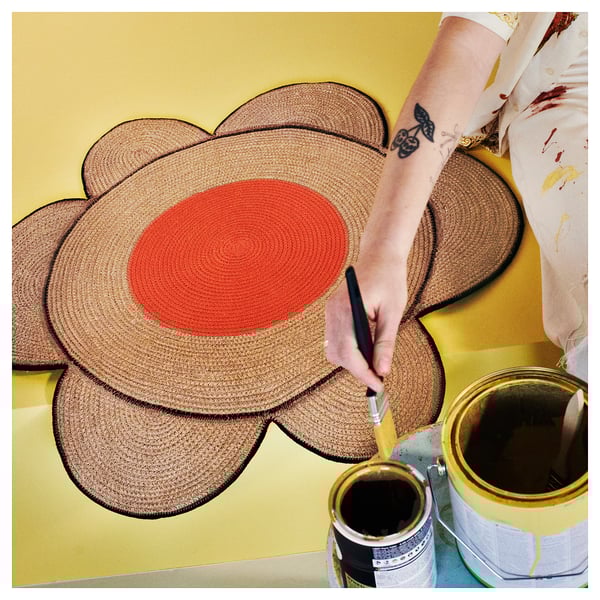 Person painting a large orange circular center on a woven brown flower-shaped rug on a yellow background.