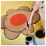 Person painting a large orange circular center on a woven brown flower-shaped rug on a yellow background.